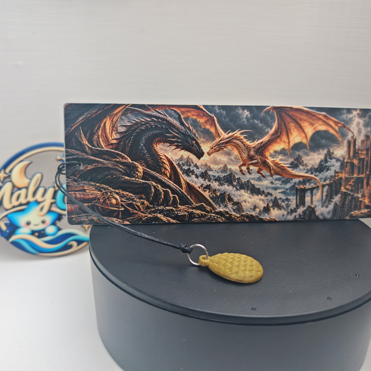 Vue rapprochée du recto du marque-page fantasy en relief, présenté sur un socle noir. Le dragon noir déploie ses ailes face à un dragon doré en vol, dans un paysage de montagnes, de brume et d’éclairs, avec une forteresse sur la droite. Le cordon noir traverse le premier plan et rejoint un anneau argenté portant une breloque œuf de dragon dorée, dont les écailles sont visibles.
Close-up of the front of the raised fantasy bookmark on a black stand. A black dragon spreads its wings opposite a flying gold drag