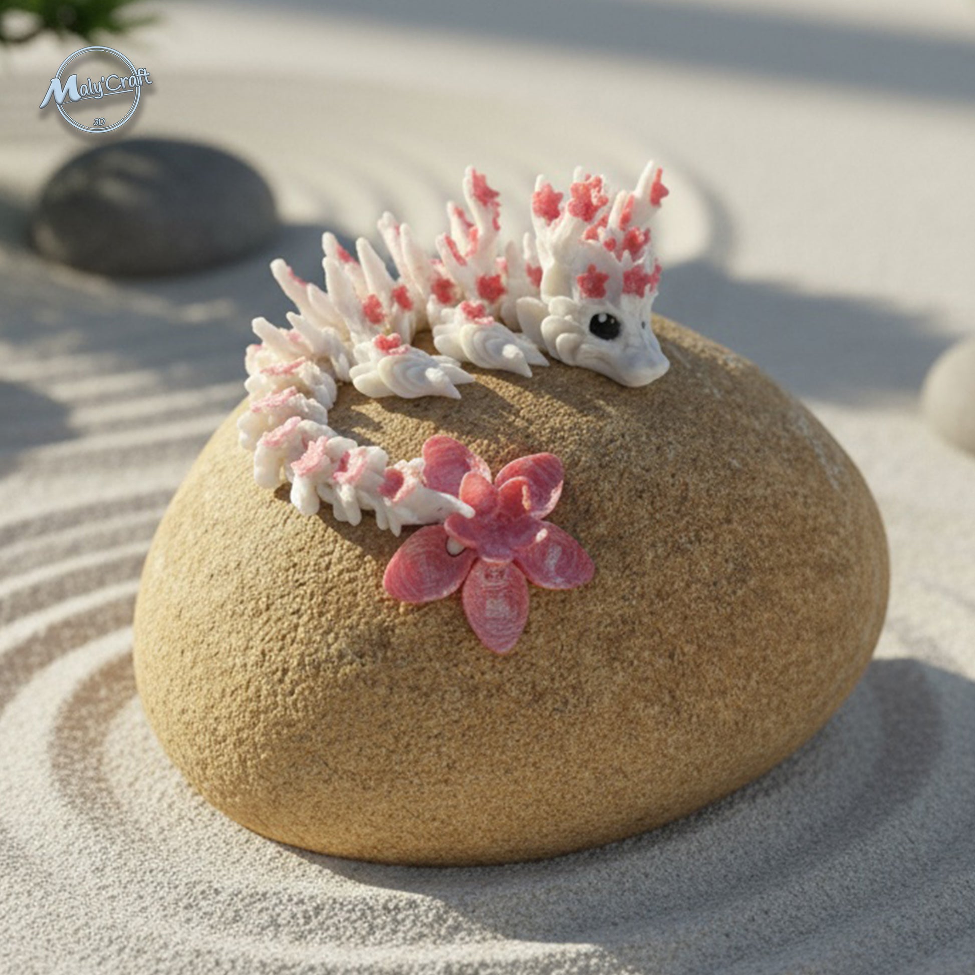 Petit dragon articulé blanc et rose à motif floral, allongé sur un galet beige dans un décor zen de sable fin. Création Cinderwings imprimée en 3D, au style délicat et poétique, parfaite pour la décoration ou la collection.