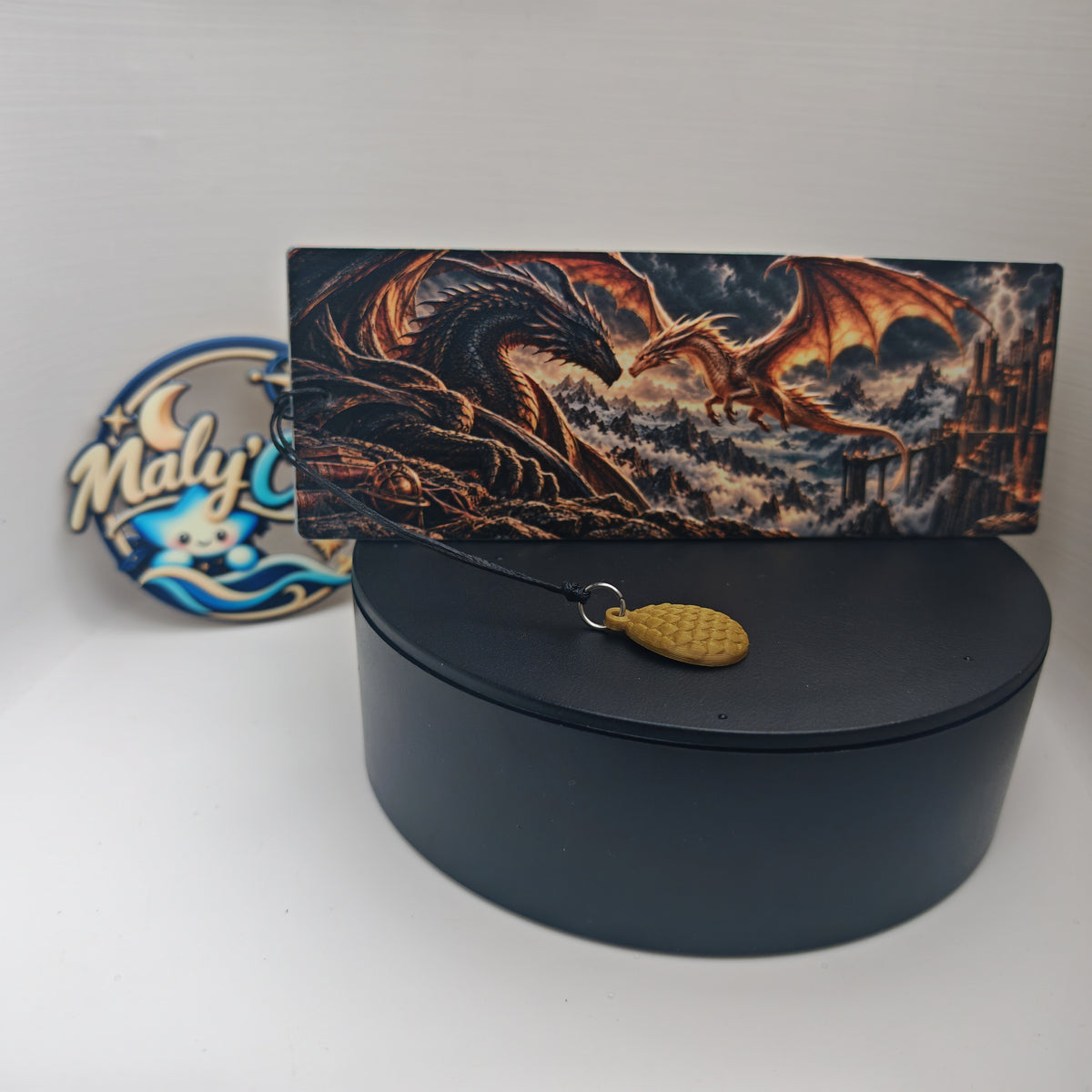 Marque-page dragon en relief présenté horizontalement sur un socle noir, sur fond clair. L’illustration représente un dragon noir et un dragon doré face à face, au-dessus de montagnes brumeuses, avec une forteresse à droite. Au premier plan, une breloque œuf de dragon dorée aux écailles en relief est attachée au cordon noir par un anneau argenté. Le logo Maly’Craft apparaît derrière le produit, à gauche.
Raised dragon bookmark displayed horizontally on a black stand against a light background. The artwork f