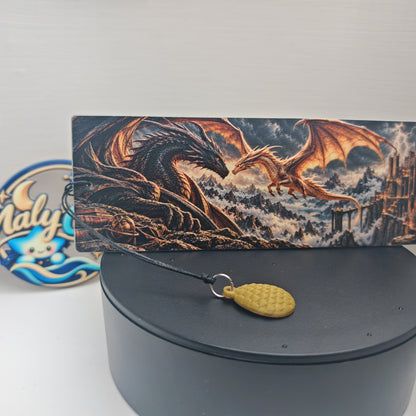 Vue rapprochée du recto du marque-page fantasy en relief, présenté sur un socle noir. Le dragon noir déploie ses ailes face à un dragon doré en vol, dans un paysage de montagnes, de brume et d’éclairs, avec une forteresse sur la droite. Le cordon noir traverse le premier plan et rejoint un anneau argenté portant une breloque œuf de dragon dorée, dont les écailles sont visibles.
Close-up of the front of the raised fantasy bookmark on a black stand. A black dragon spreads its wings opposite a flying gold drag