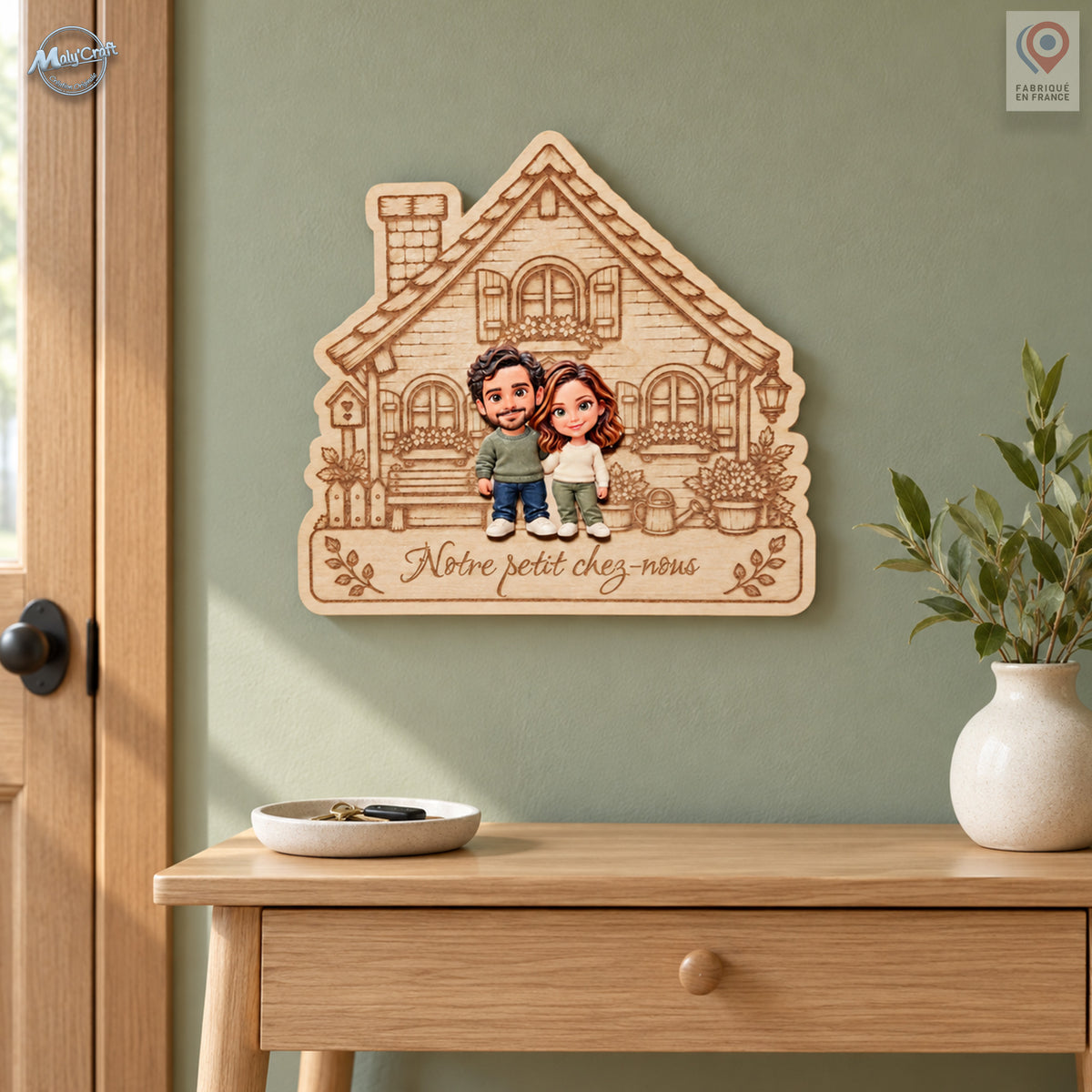 Mise en situation d’une maison en bois gravé personnalisée pour un couple, portant l’inscription Notre petit chez-nous. La décoration est accrochée sur un mur vert sauge près d’une porte en bois. Une console en chêne, une coupelle à clés et un vase écru avec des branches feuillues complètent cette scène d’entrée.
Lifestyle view of a personalized wooden couple house plaque engraved with the French wording Notre petit chez-nous. It hangs on a sage-green wall beside a wooden entrance door, above an oak console