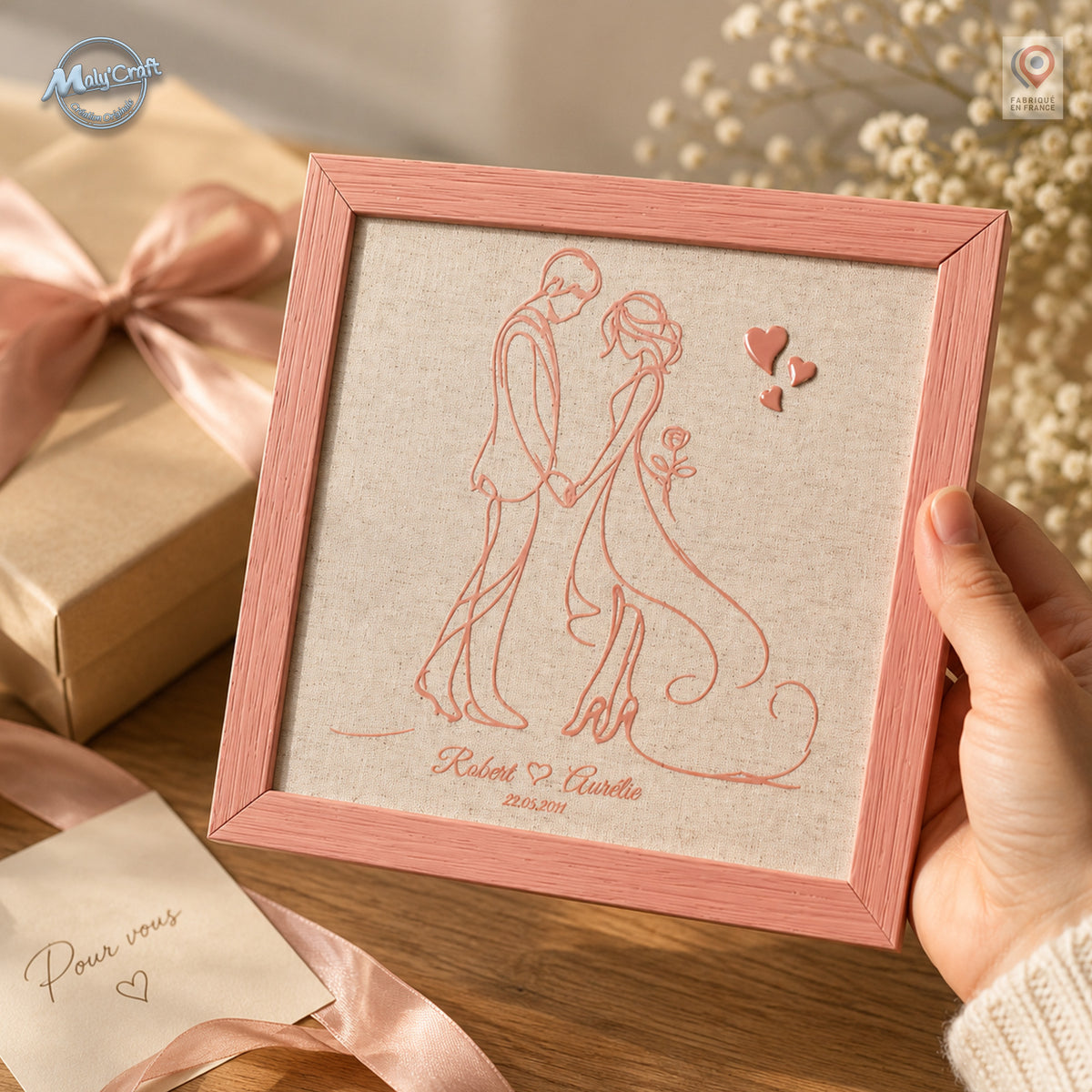 Cadre de mariage personnalisé tenu en main, avec portrait de couple en relief sur lin, prénoms et date. Mise en scène cadeau avec boîte, ruban rose et fleurs dans une ambiance douce et romantique.
Personalized wedding frame held in hand, featuring a raised couple portrait on linen with custom names and date. Gift setting with a box, pink ribbon and flowers in a soft romantic atmosphere.