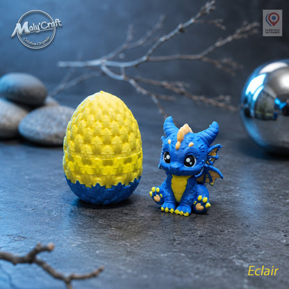 Figurine bébé dragon Éclair bleu électrique et jaune avec son œuf bicolore jaune et bleu, posés sur une surface en ardoise sombre. Création originale MalyCraft3D, impression 3D de précision réalisée en France. Dragon kawaii multicolore avec détails écailles et cornes dorées. Ambiance électrique avec galets, branches sèches et sphère chromée en arrière-plan.