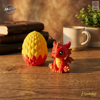 Figurine bébé dragon Flamme rouge et jaune avec son œuf bicolore jaune et rouge, posés sur une table en bois. Création originale MalyCraft3D, impression 3D de précision réalisée en France. Dragon kawaii multicolore avec détails écailles et cornes dorées. Œuf en deux parties emboîtables. Ambiance cosy avec livres anciens, tasse blanche et plante verte en arrière-plan.