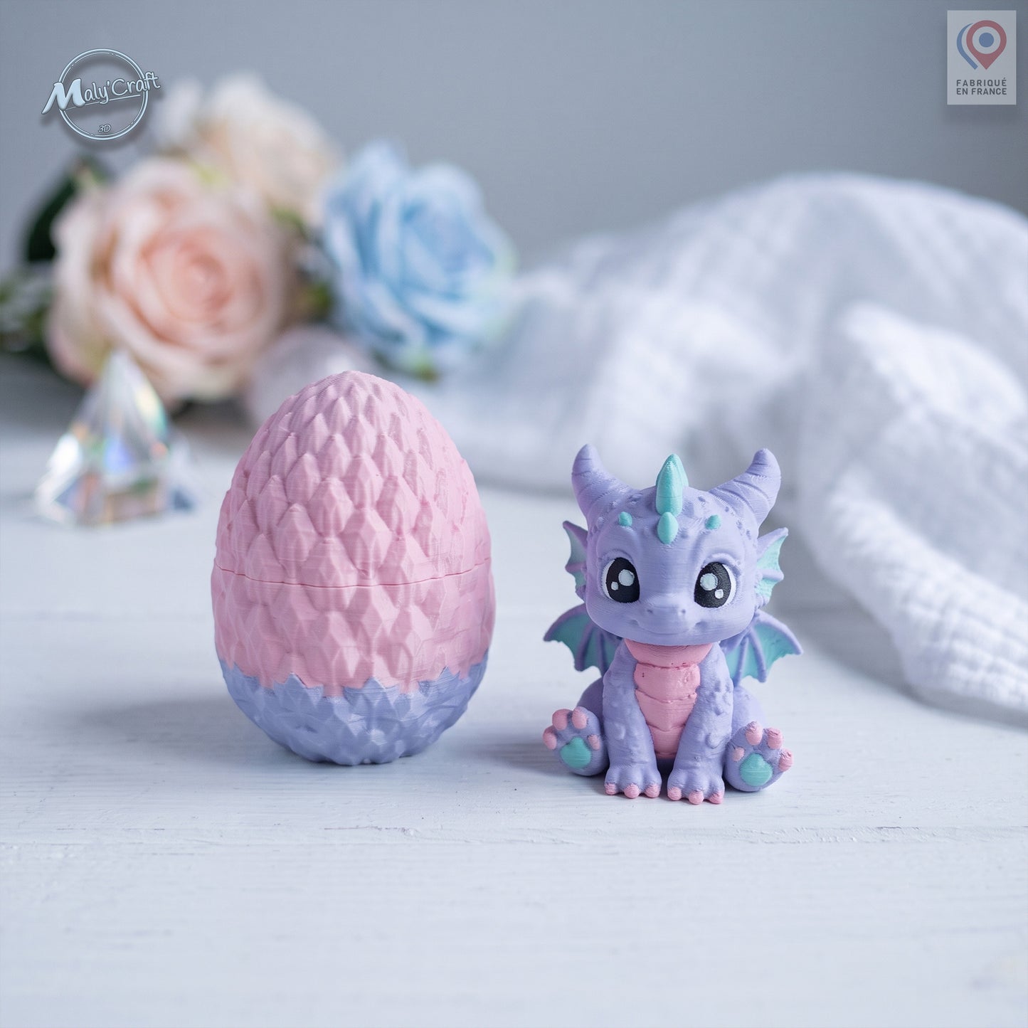 Figurine bébé dragon Licorne violet et rose avec son œuf bicolore rose et violet, posés sur une surface blanche. Création originale MalyCraft3D, impression 3D de précision réalisée en France. Dragon kawaii multicolore avec détails écailles et cornes turquoise. Ambiance féérique avec fleurs pastel roses et bleues, cristal et tissu blanc en arrière-plan.