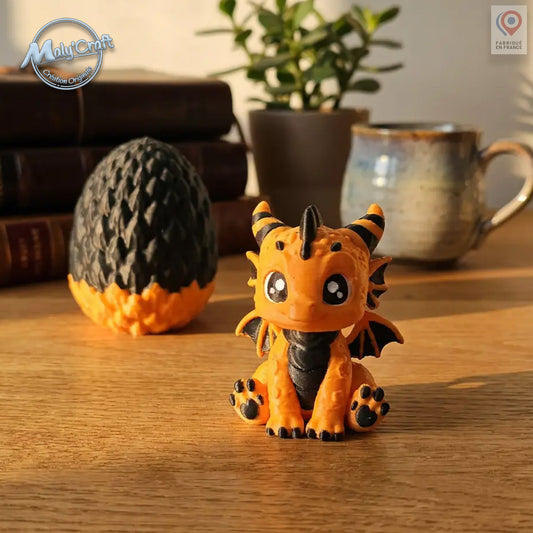 Figurine bébé dragon Tigre orange et noir avec son œuf bicolore noir et orange, posés sur une table en bois. Création originale MalyCraft3D, impression 3D de précision réalisée en France. Dragon kawaii multicolore avec détails écailles et cornes rayées. Œuf en deux parties emboîtables. Ambiance cosy avec livres anciens, tasse céramique et plante verte en arrière-plan.