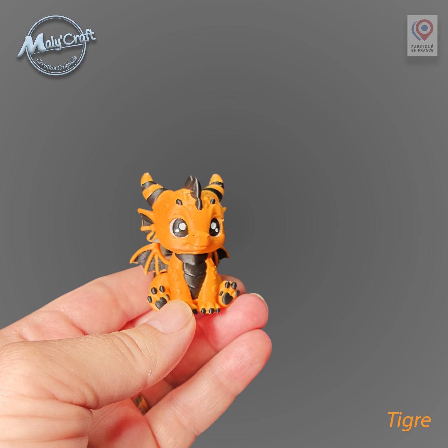 Main tenant la figurine bébé dragon Tigre orange et noir pour montrer sa taille miniature. Création originale MalyCraft3D, impression 3D de précision réalisée en France. Dragon kawaii multicolore avec cornes rayées orange et noir, détails écailles visibles. Fond gris anthracite avec logo MalyCraft3D et badge Fabriqué en France.