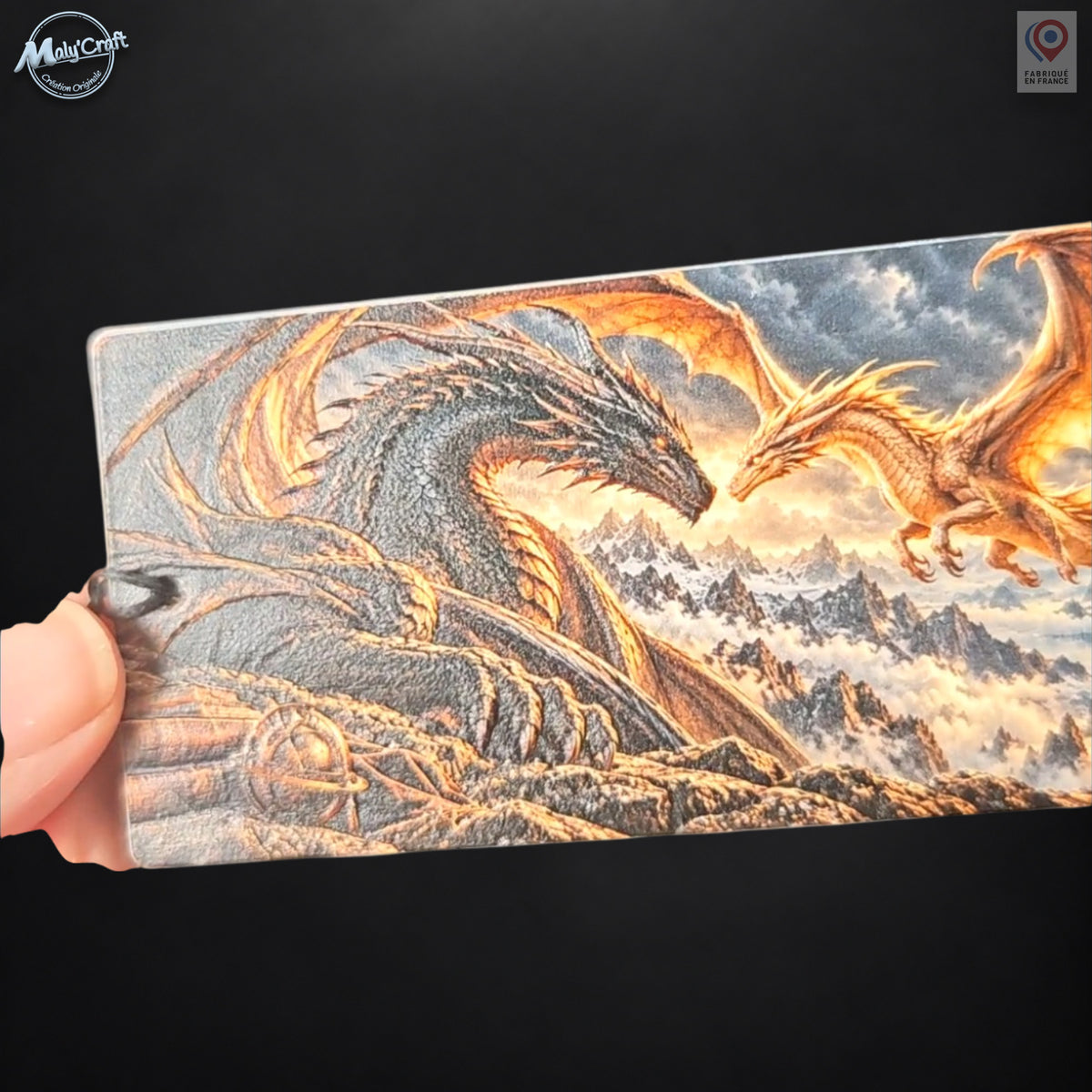 Gros plan du recto du marque-page dragon, tenu par son bord gauche devant un fond sombre. Les reflets mettent en évidence la texture de surface et les reliefs de l’illustration représentant un dragon noir face à un dragon doré. Le cadrage rapproché privilégie les détails du motif. Le logo Maly’Craft figure dans le coin supérieur gauche.
Close-up of the front of the dragon bookmark, held at its left edge against a dark background. Reflections reveal the surface texture and raised details of the artwork featu