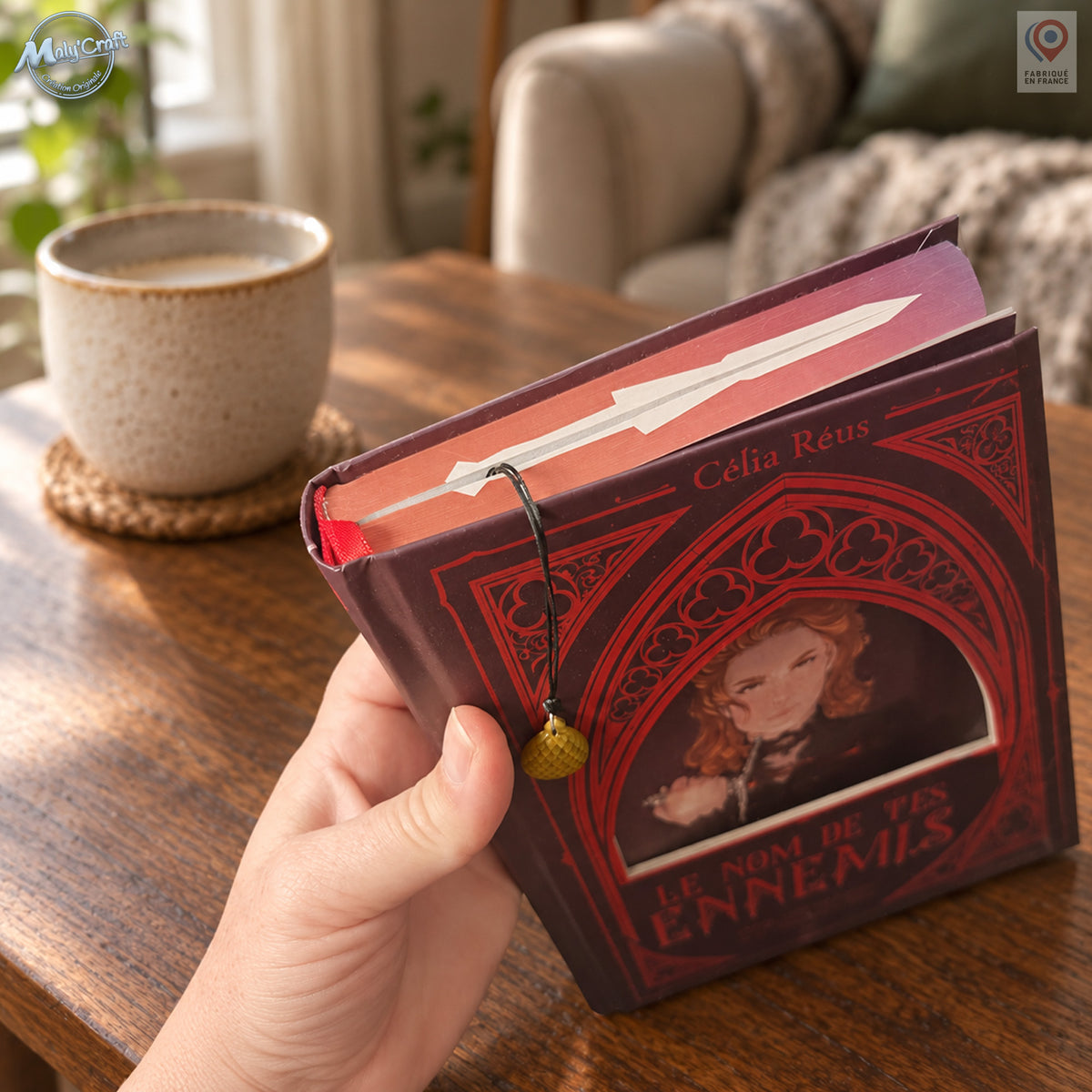 Vue du marque-page utilisé dans un livre fermé tenu à la main. Le cordon noir passe sur la tranche supérieure du livre et laisse pendre une breloque œuf de dragon dorée devant la couverture rouge illustrée. Le décor reprend le coin lecture avec une table en bois, une tasse en céramique et un fauteuil beige en arrière-plan.
Bookmark in use inside a closed book held in one hand. The black cord passes over the top page edge, leaving a gold-coloured dragon egg charm hanging against the illustrated red cover. A 