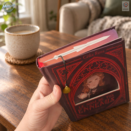 Vue du marque-page utilisé dans un livre fermé tenu à la main. Le cordon noir passe sur la tranche supérieure du livre et laisse pendre une breloque œuf de dragon dorée devant la couverture rouge illustrée. Le décor reprend le coin lecture avec une table en bois, une tasse en céramique et un fauteuil beige en arrière-plan.
Bookmark in use inside a closed book held in one hand. The black cord passes over the top page edge, leaving a gold-coloured dragon egg charm hanging against the illustrated red cover. A 