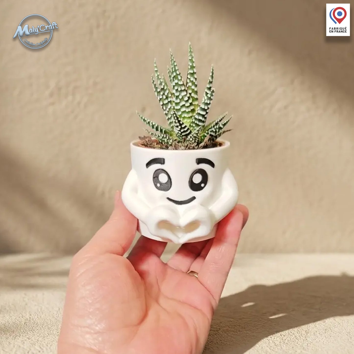 Un petit pot de fleurs blanc en forme de personnage stylisé avec des mains formant un cœur à sa base. Le pot contient une petite plante succulente (Aloe Vera) et présente un visage souriant aux grands yeux expressifs. L'objet est une impression 3D de précision présentée à la main sur un fond neutre ensoleillé.