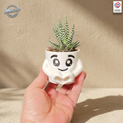 Un petit pot de fleurs blanc en forme de personnage stylisé avec des mains formant un cœur à sa base. Le pot contient une petite plante succulente (Aloe Vera) et présente un visage souriant aux grands yeux expressifs. L'objet est une impression 3D de précision présentée à la main sur un fond neutre ensoleillé.