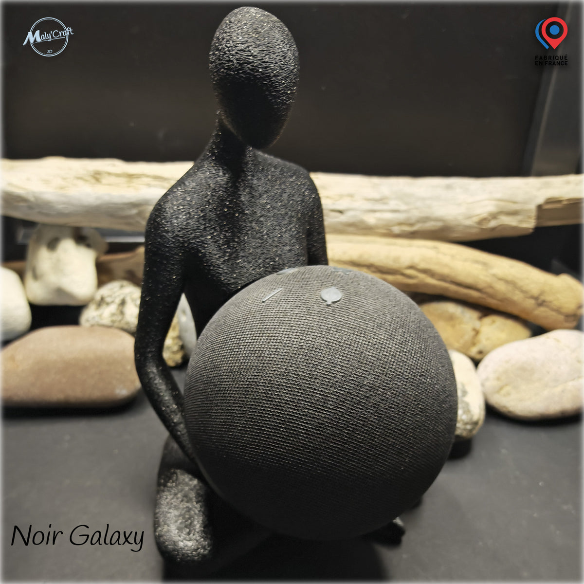 Photographie réelle d'un support décoratif pour enceinte connectée Amazon Echo Dot Alexa, fabriqué en France par MalyCraft. La figurine abstraite sans visage, imprimée en 3D dans un filament noir Galaxy légèrement scintillant, représente un personnage assis tenant une enceinte sphérique grise entre ses bras. La finition texturée de la figurine contraste avec le revêtement textile de l'enceinte, dont les boutons de commande sont visibles sur le dessus. L'ensemble est photographié devant un décor composé de g