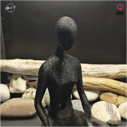 Photographie rapprochée du support décoratif pour enceinte connectée Amazon Echo Dot Alexa, fabriqué en France par MalyCraft. Cette figurine abstraite imprimée en 3D représente un personnage sans visage en position assise, avec une finition noir Galaxy légèrement scintillante. Le cadrage met en évidence la texture réelle de l'impression 3D, notamment les reliefs visibles sur la tête, les épaules, le buste et les bras. Présenté sans enceinte devant un décor de galets naturels et de bois flotté, ce gros plan 