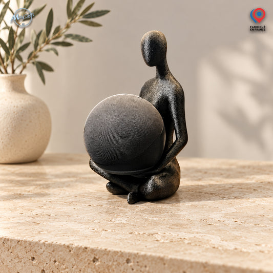 Support décoratif pour enceinte connectée Amazon Echo Dot Alexa, composé d'une figurine abstraite sans visage, assise et tenant une enceinte sphérique grise entre ses bras. Réalisée en impression 3D avec une finition noire texturée, la figurine repose directement sur une surface en pierre beige clair, dans un décor contemporain avec un vase en céramique et des branches d'olivier. Le logo MalyCraft apparaît en haut à gauche et le pictogramme Fabriqué en France en haut à droite. Un accessoire design original 
