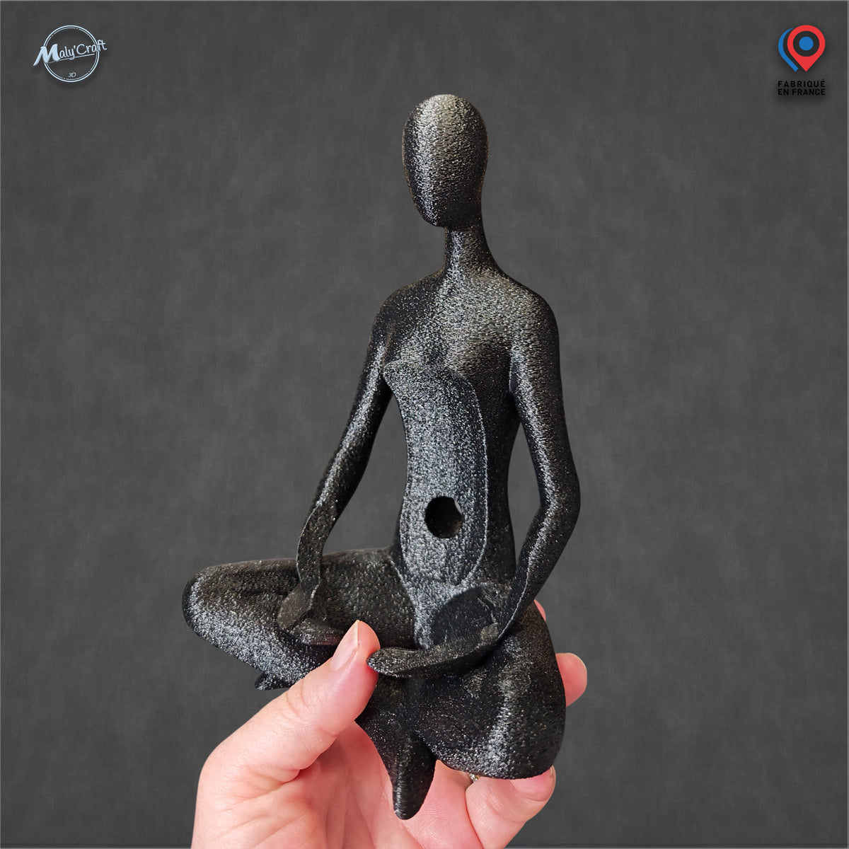 Vue détaillée du support décoratif pour enceinte connectée Amazon Echo Dot Alexa, photographié sans enceinte afin de montrer sa conception et sa finition. La figurine abstraite sans visage, imprimée en 3D dans un filament noir Galaxy légèrement scintillant, représente un personnage assis en tailleur, les bras positionnés pour accueillir une enceinte sphérique. Une ouverture circulaire est visible au centre du support. La figurine est tenue dans une main devant un fond gris anthracite, permettant d'apprécier